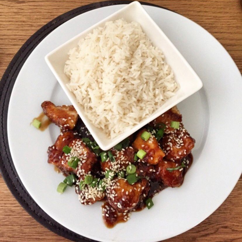 A plate of glazed chicken and sesame seeds next to a bowl of white rice. 